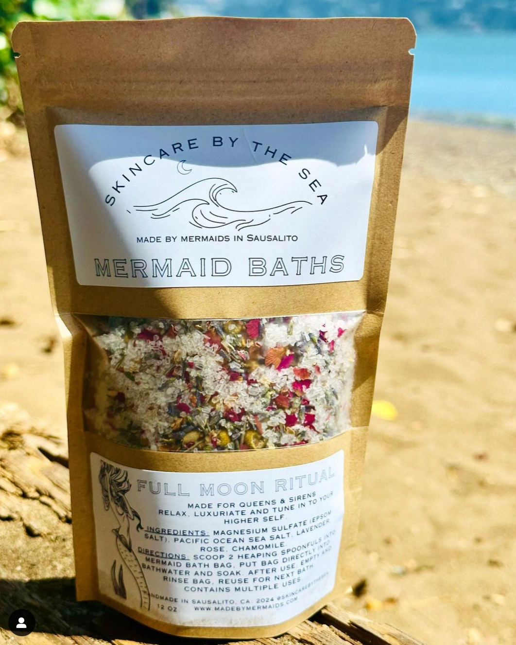 A sealed pouch of Mermaid Baths brand 'Full Moon Ritual' bath salt blend, featuring Epsom salt, Pacific Ocean sea salt, rose, lavender, and chamomile flowers, displayed on a wooden surface with a bright background.