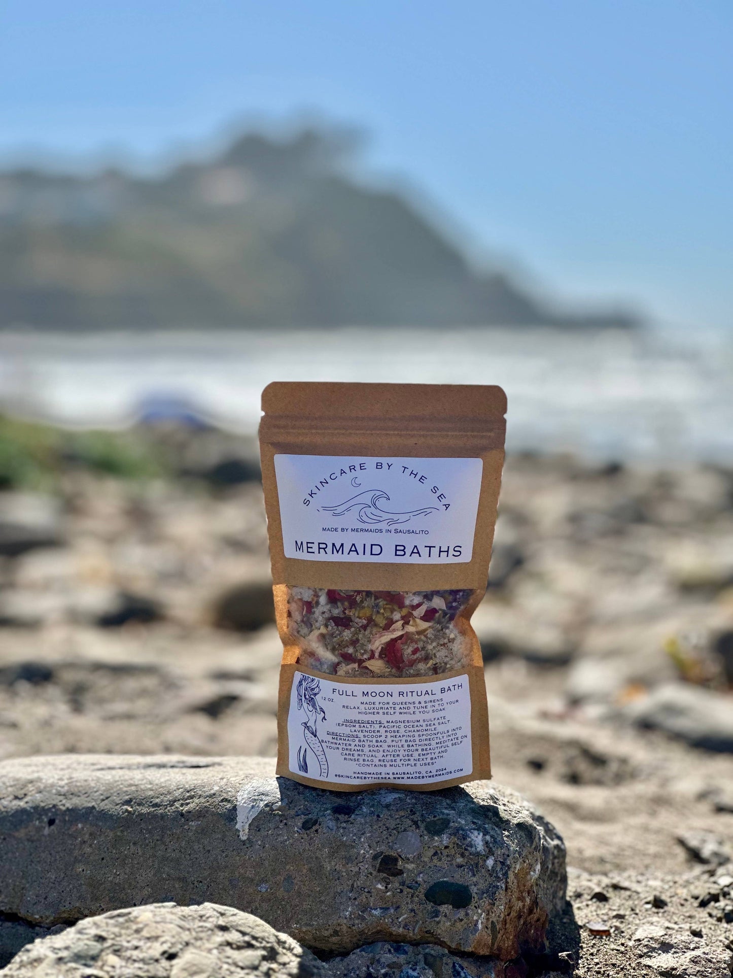 A product image of a package labeled 'Mermaid Bath: Full Moon Ritual' with a mix of bath salts and flowers visible through the packaging, placed on a rock with a blurred background of a beach setting.