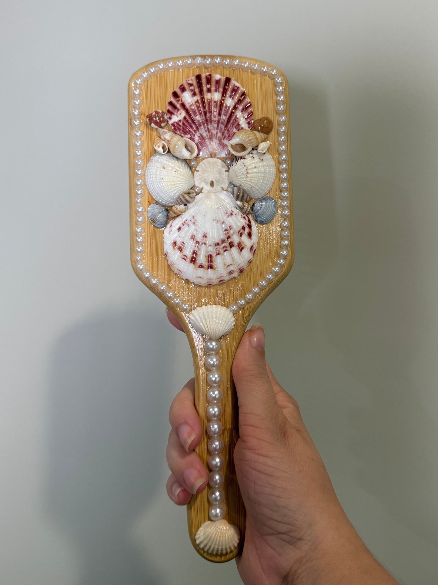 Hand holding a wooden hair brush with seashells against a plain background