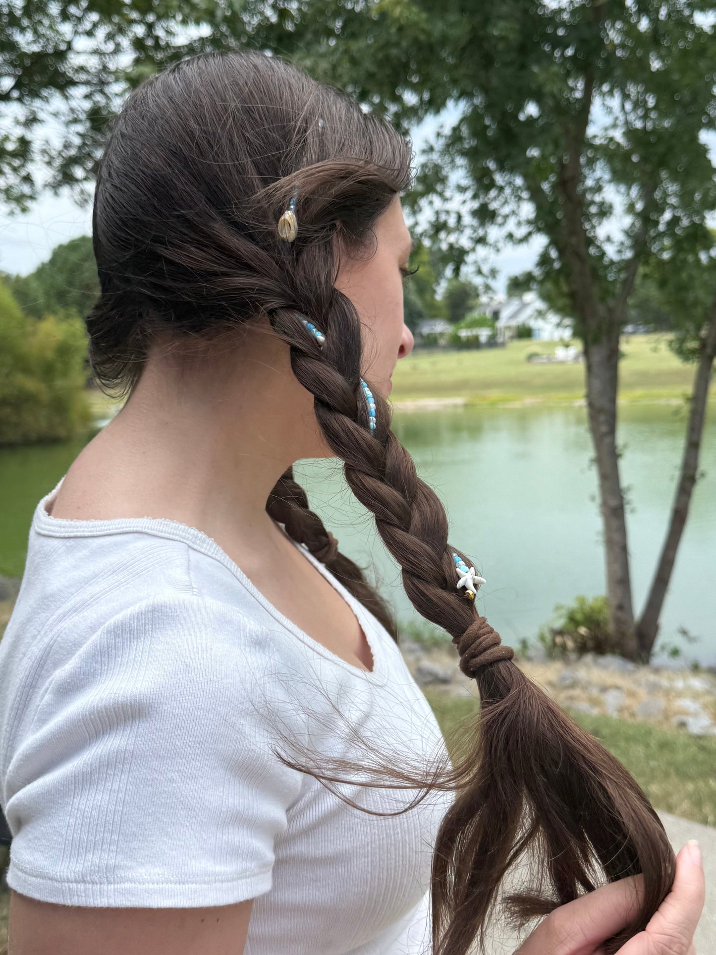 Person with braided hair a seashell hair extension clip standing outdoors near a body of water