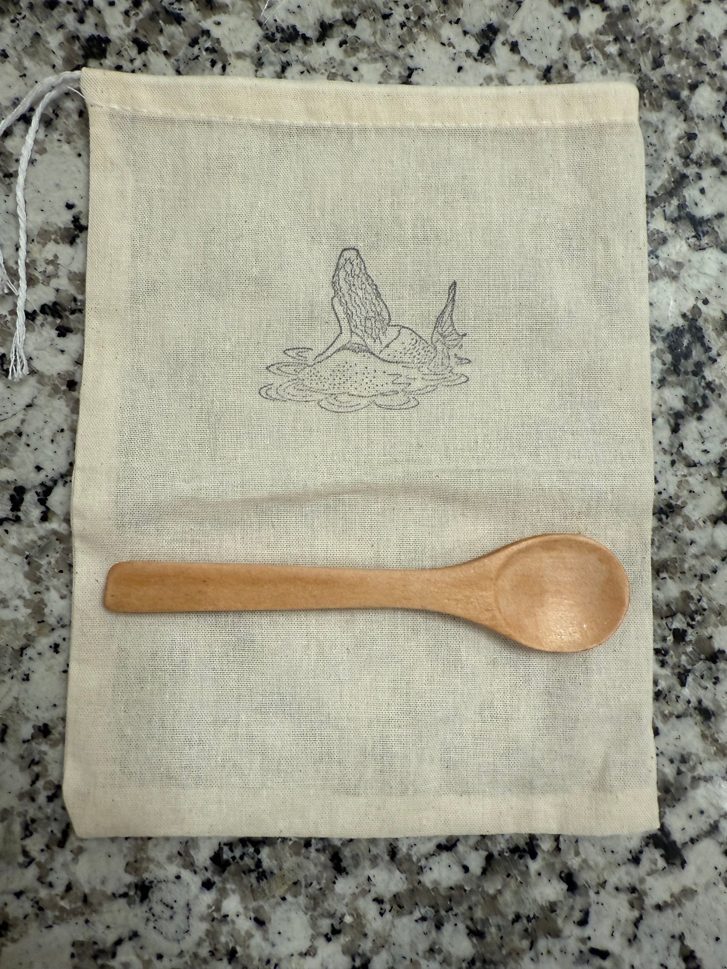 Wooden spoon on a fabric pouch with a mermaid design showing that the bath salts come with a reusable pouch.
