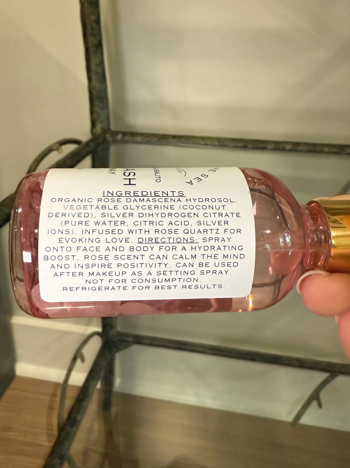 Hand holding a bottle of 'Siren Splash' showing the ingredients Organic Rose Damascena Hydrosol, Vegetable Glycerin (Coconut Deprived), Silver Dihydrogen Citrate (Pure water, citric acid, silver ions) Rose Quartz Crystals.