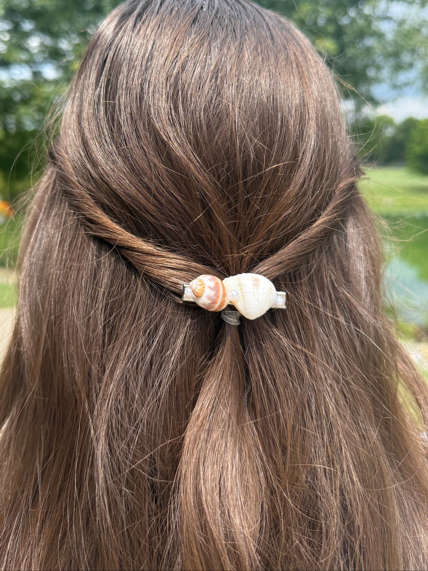 Close-up of a person's hair with a decorative seashell hair clip against a blurred green outdoor background