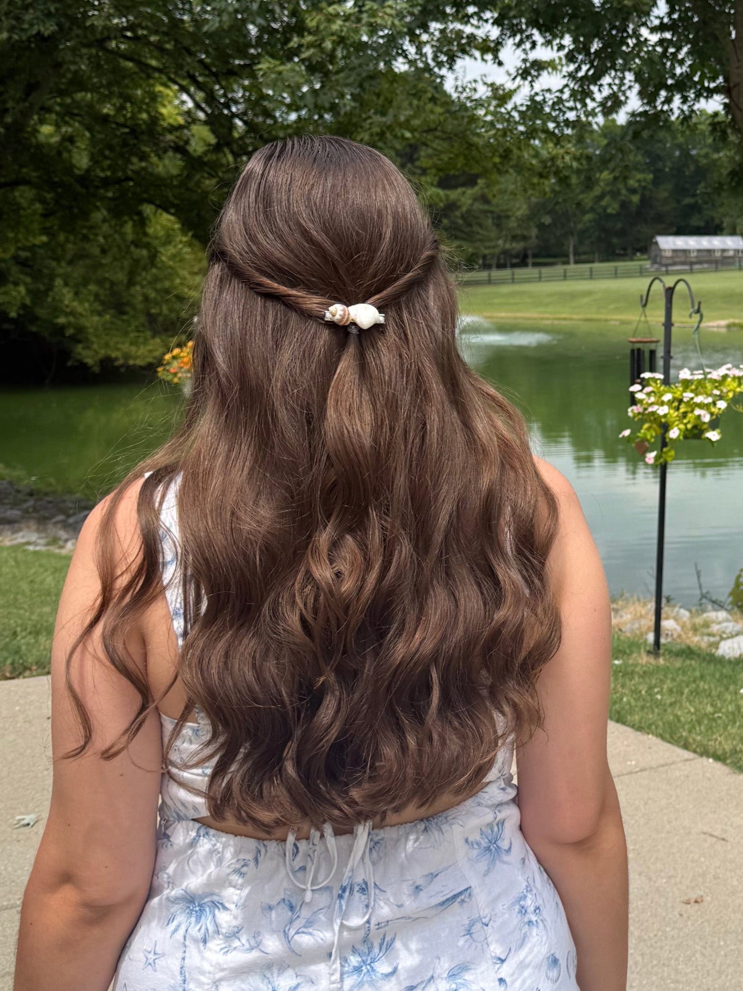 Person with long brown hair wearing a seashell hair clip standing by a pond with trees and grass in the background