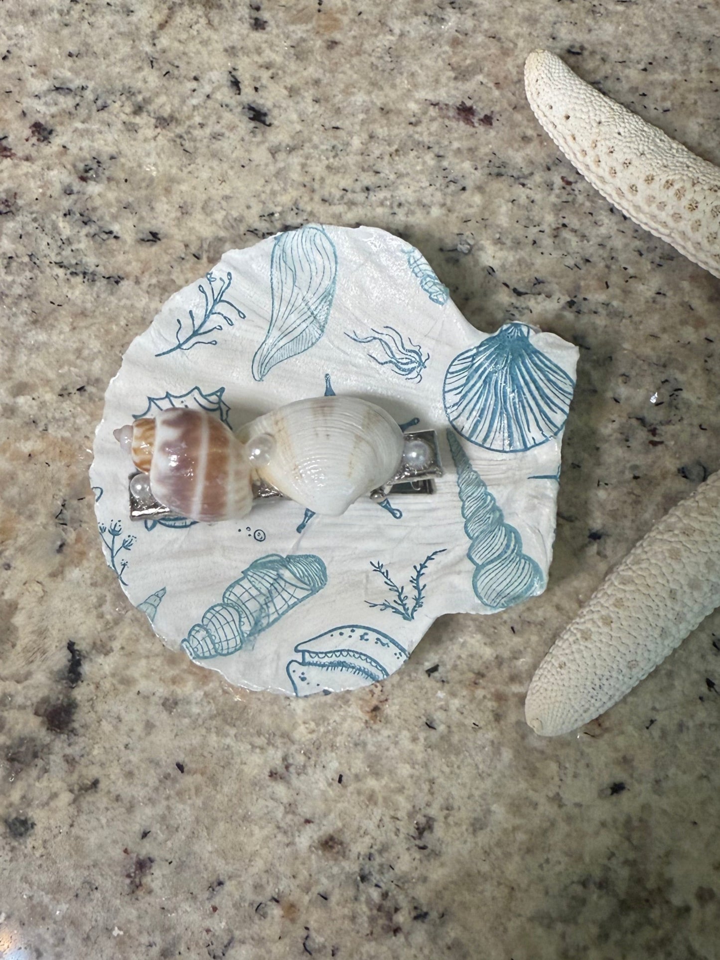 Seashell hair clip sitting in a seashell trinket dish on a beige background
