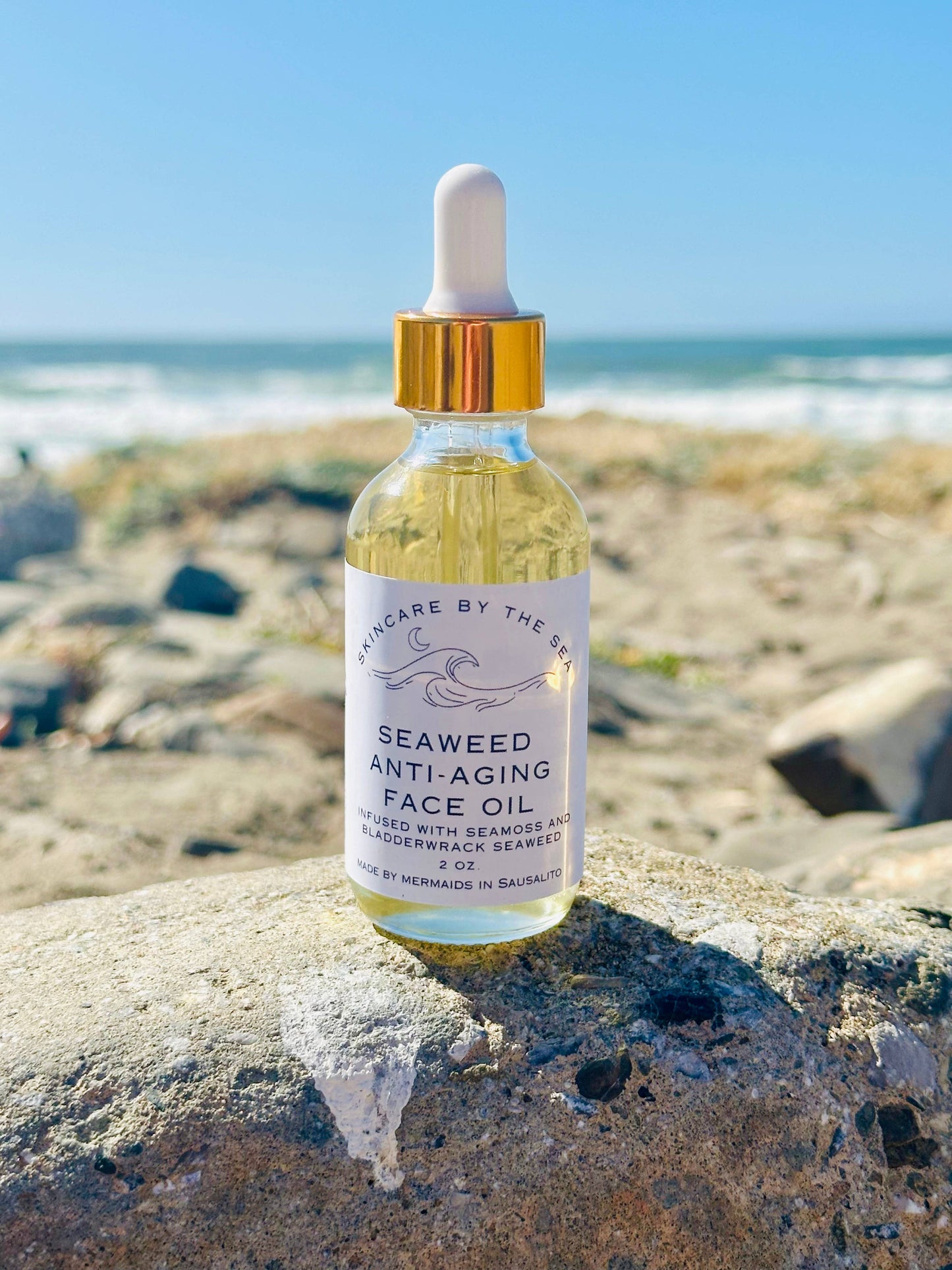 A bottle of Seaweed Anti-Aging Face Oil with a dropper, placed on a rock at the beach, with the ocean and sky in the background.