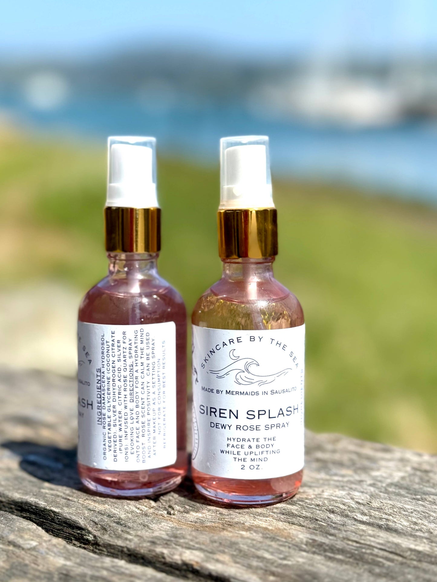 Two bottles of 'Siren Splash' dewy rose spray on a wooden surface with a blurred natural background.
