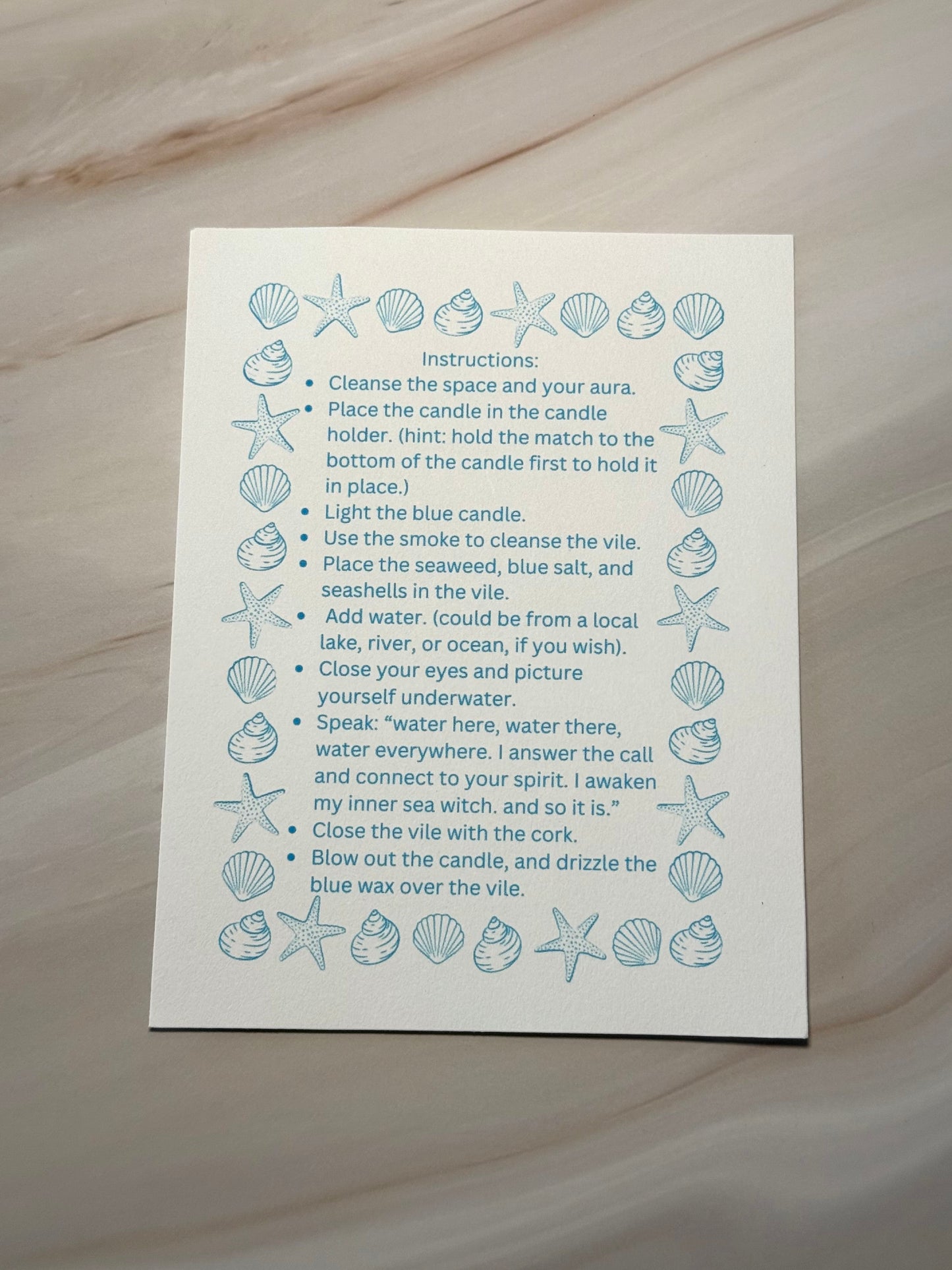 Instructions for a ritual on a white card with blue illustrations of seashells and starfish, placed on a textured beige surface.