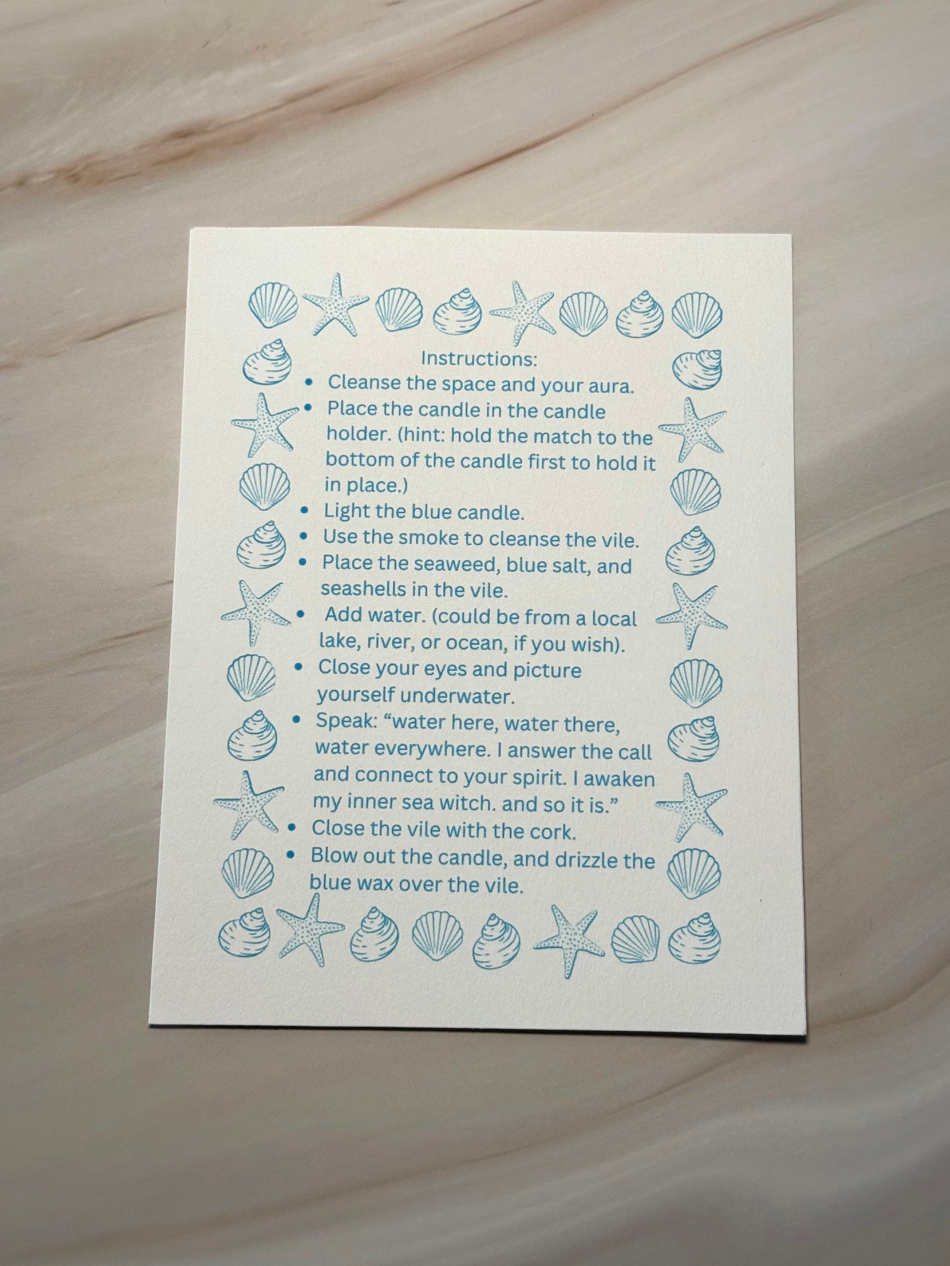Instructions for a ritual on a white card with blue illustrations of seashells and starfish, placed on a textured beige surface.