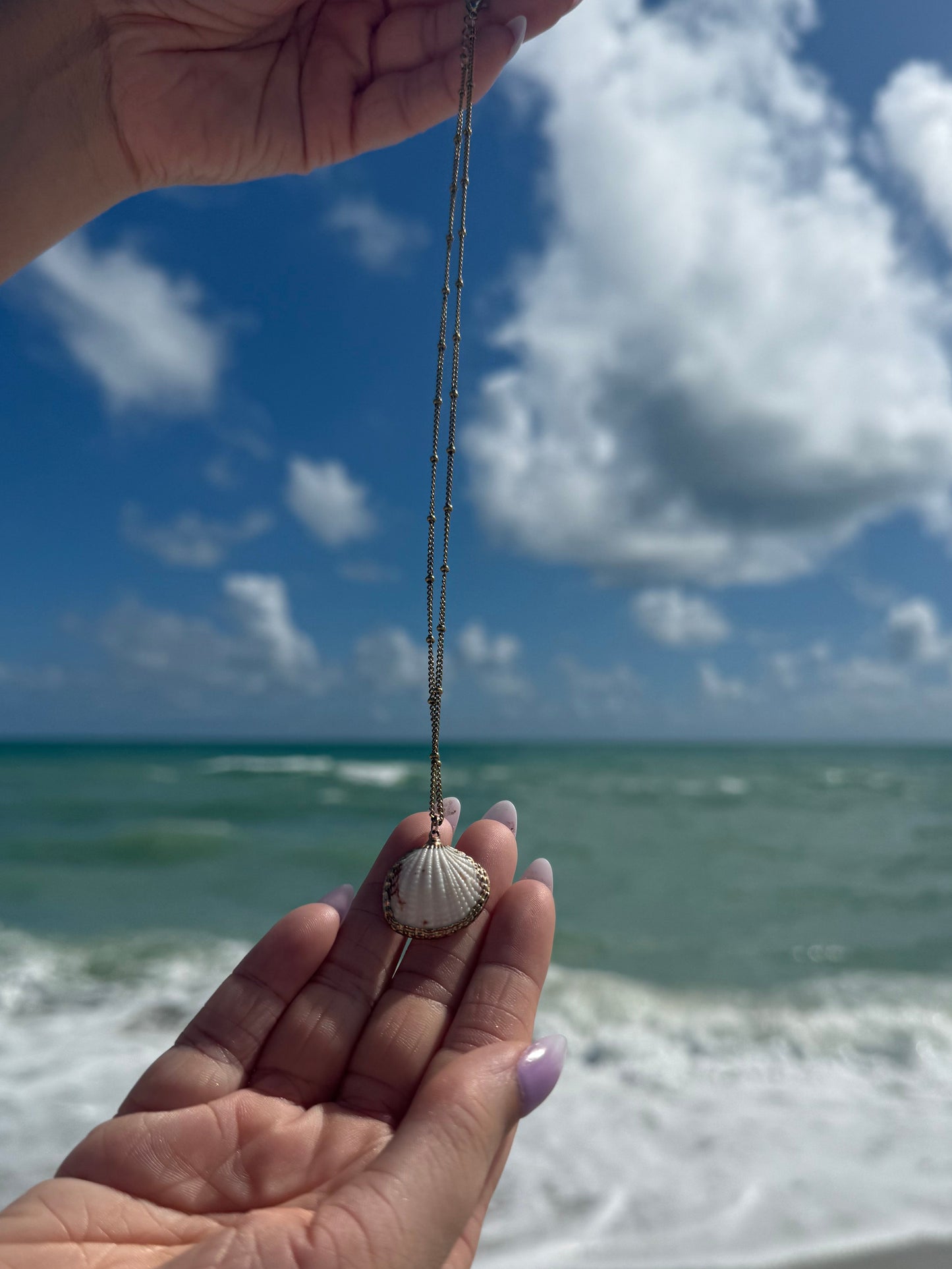 Person holding a shell necklace with a gold chain against a blue sky and white wall.