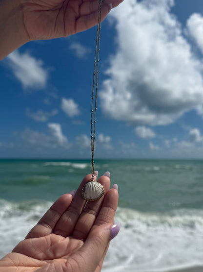 Person holding a shell necklace with a gold chain against a blue sky and white wall.