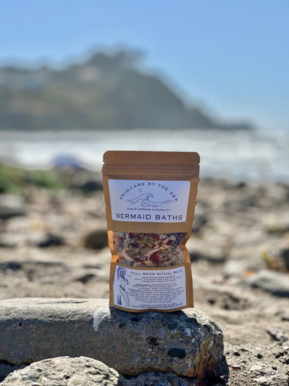 A product image of a package labeled 'Mermaid Bath: Full Moon Ritual' with a mix of bath salts and flowers visible through the packaging, placed on a rock with a blurred background of a beach setting.