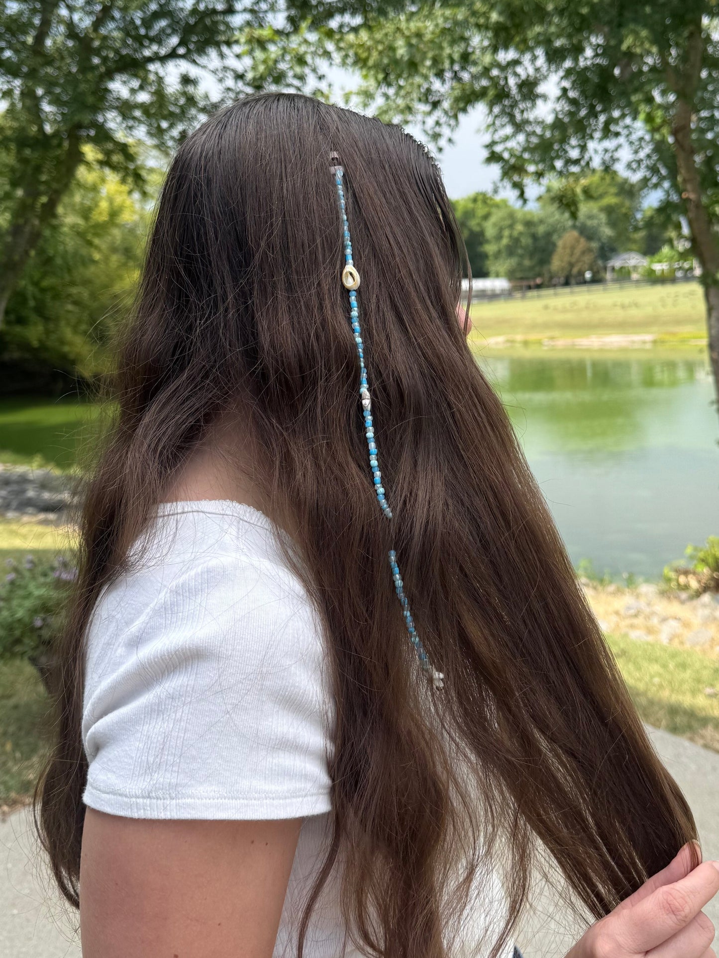 Person with long hair featuring a decorative seashell hair extension clup, standing outdoors near a body of water.