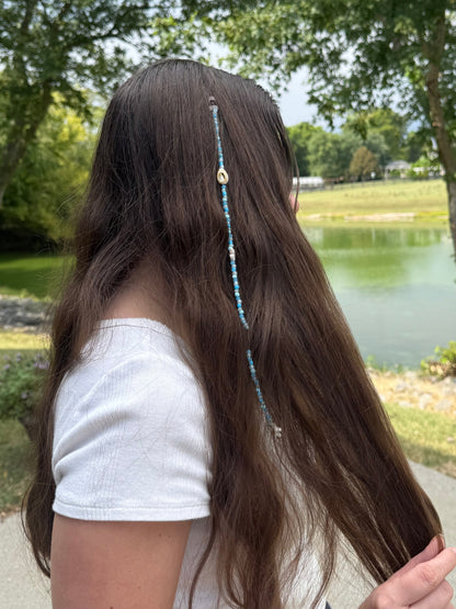 Person with long hair featuring a decorative seashell hair extension clup, standing outdoors near a body of water.