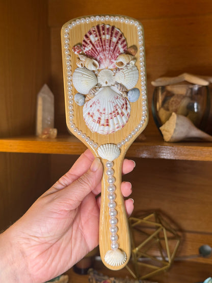 Hand holding a decorative brush with shell designs in front of a wooden cabinet.