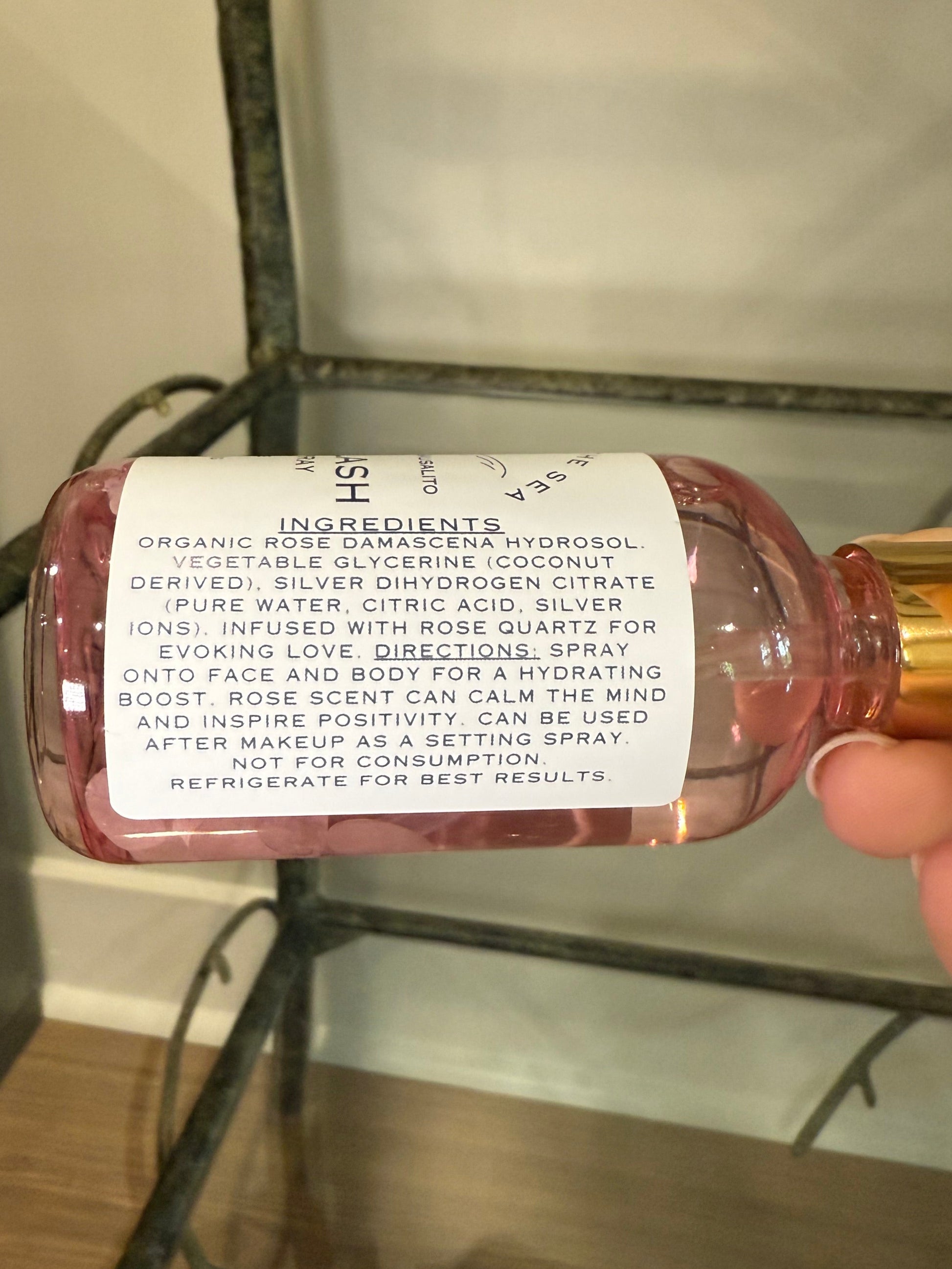 Hand holding a bottle of 'Siren Splash' showing the ingredients Organic Rose Damascena Hydrosol, Vegetable Glycerin (Coconut Deprived), Silver Dihydrogen Citrate (Pure water, citric acid, silver ions) Rose Quartz Crystals.