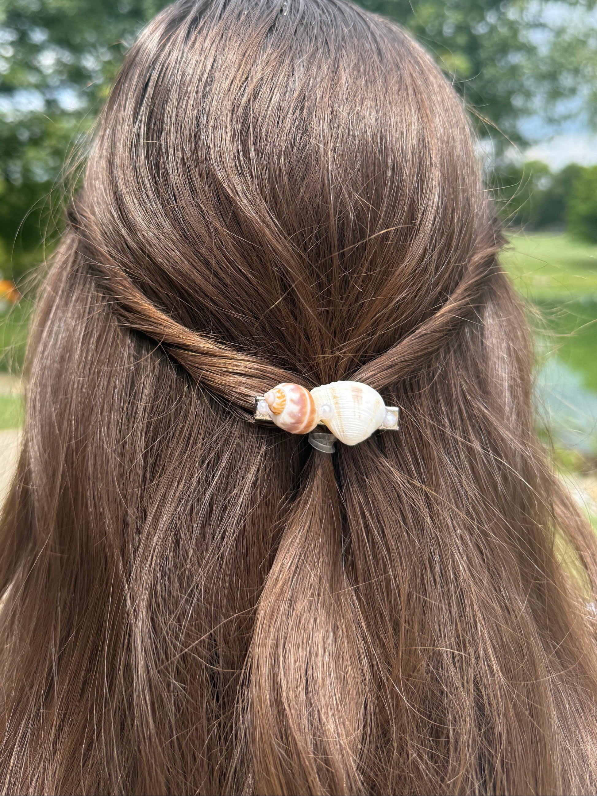 Close-up of a person's hair with a decorative seashell hair clip against a blurred green outdoor background