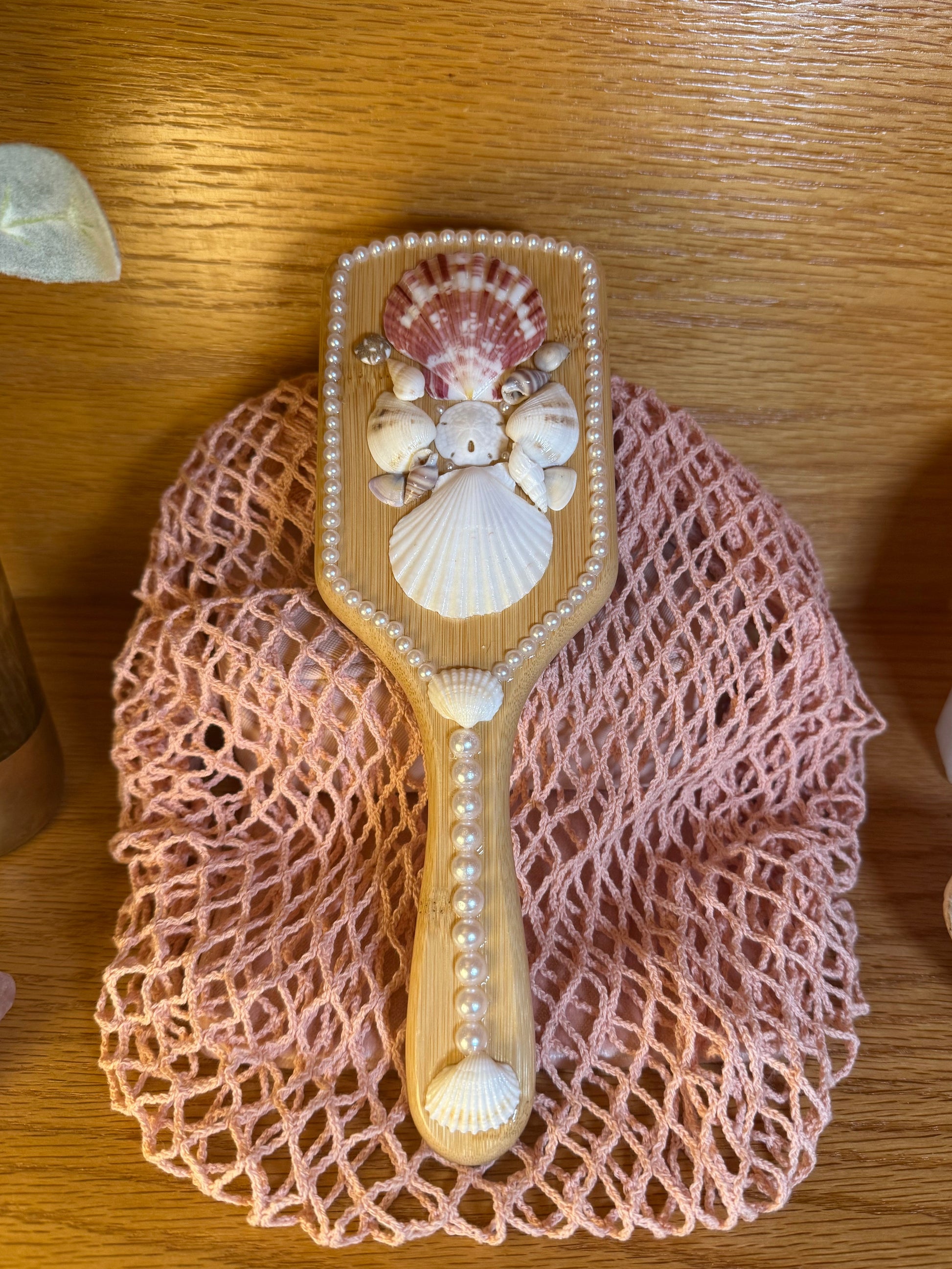 Decorative hairbrush with shell designs on a pink mesh bag on a wooden surface.