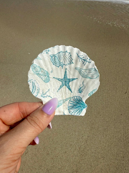 Hand holding a decorative seashell with beach-themed designs on a sandy background