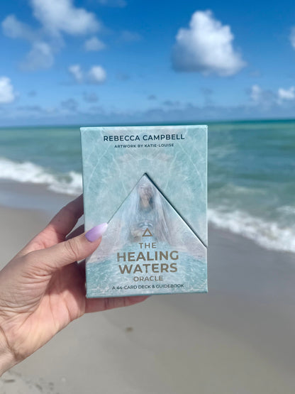 Hand holding a book titled 'The Healing Waters' by Rebecca Campbell on a beach background