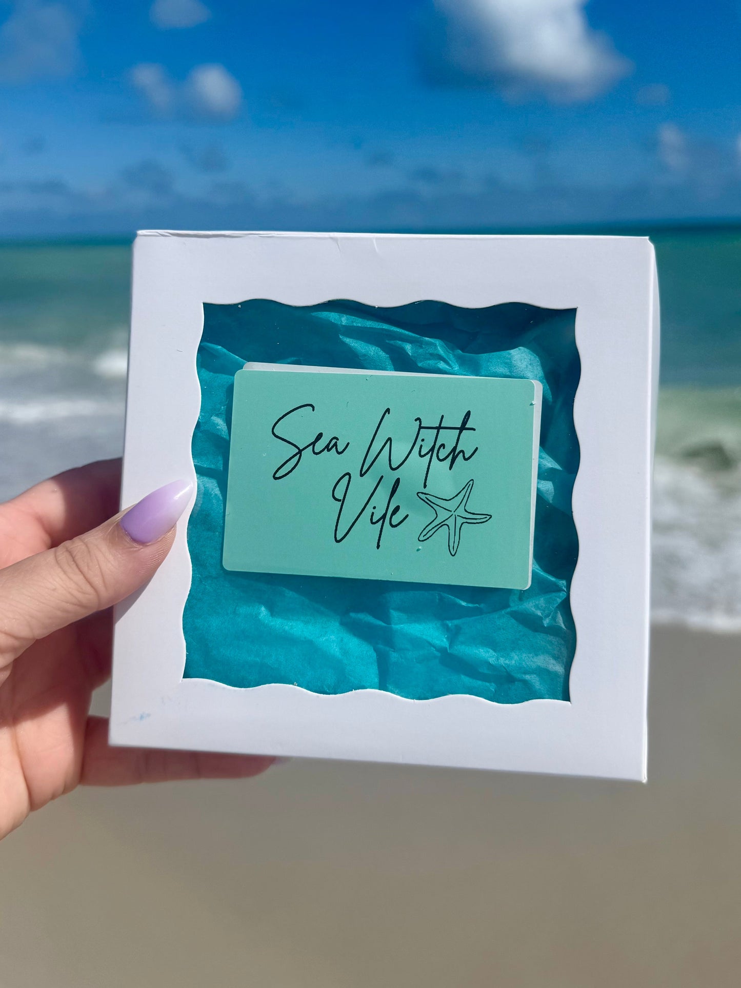 hand holding a sea witch vile spell box on a beach with sky sand and ocean background