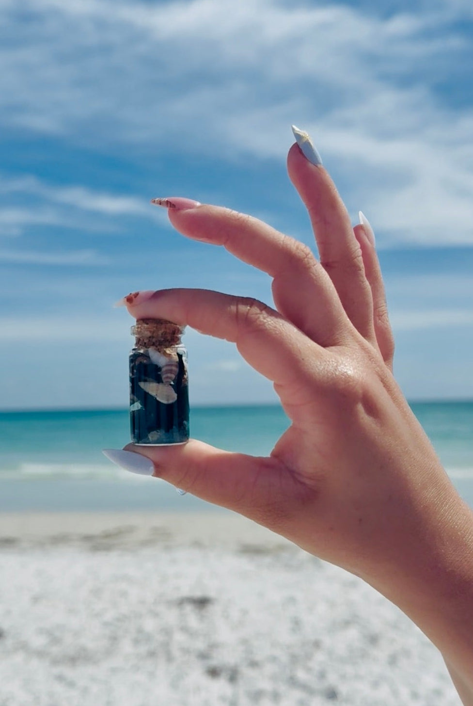 hand holding Sea witch vile bottle with seaweed and seashells on a beach with blue sky, water, and sand