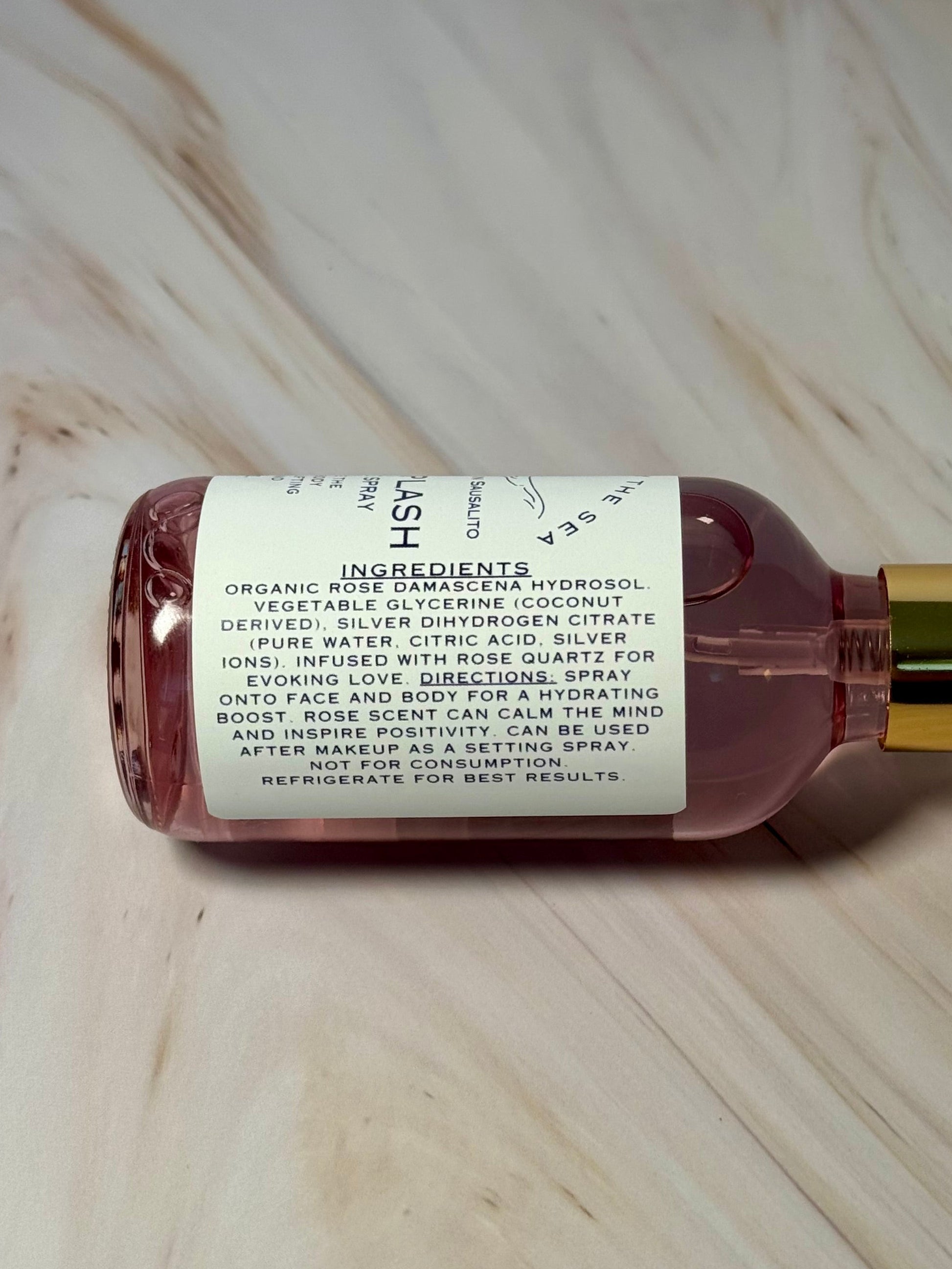 Hand holding a bottle of 'Siren Splash' showing the ingredients Organic Rose Damascena Hydrosol, Vegetable Glycerin (Coconut Deprived), Silver Dihydrogen Citrate (Pure water, citric acid, silver ions) Rose Quartz Crystals.