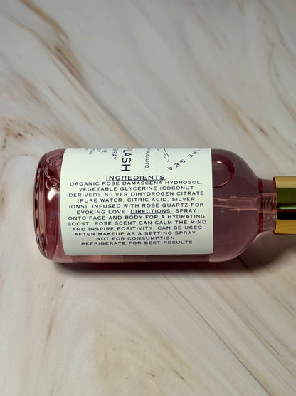 Hand holding a bottle of 'Siren Splash' showing the ingredients Organic Rose Damascena Hydrosol, Vegetable Glycerin (Coconut Deprived), Silver Dihydrogen Citrate (Pure water, citric acid, silver ions) Rose Quartz Crystals.