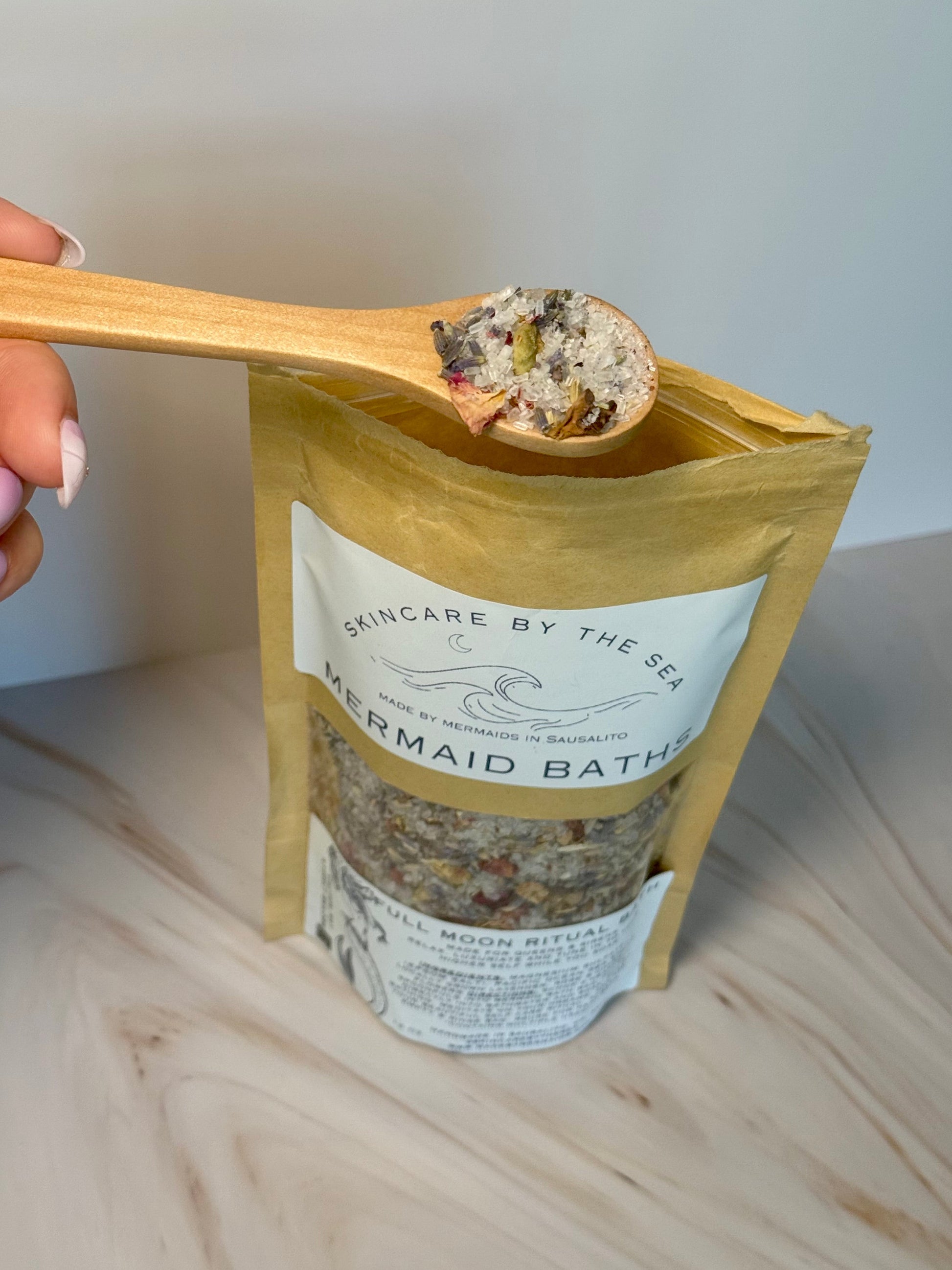 Hand holding a wooden spoon with bath salts over a package labeled 'Mermaid Bath' on a neutral background.